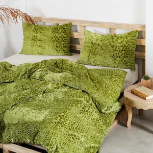 May include: A bed is made with a green velvet comforter and two matching pillowcases. The bedding has a textured pattern. The bed frame is made of light-colored wood. A small wooden side table holds a book and a plant.