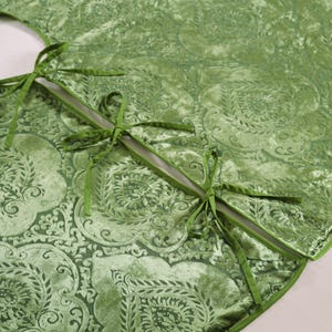 Puede incluir: Una falda de &aacute;rbol de Navidad de terciopelo verde con un patr&oacute;n floral detallado. La falda tiene un borde festoneado y se asegura con lazos de cinta verde. La tela tiene una textura suave y afelpada, y el dise&ntilde;o general es elegante y festivo.