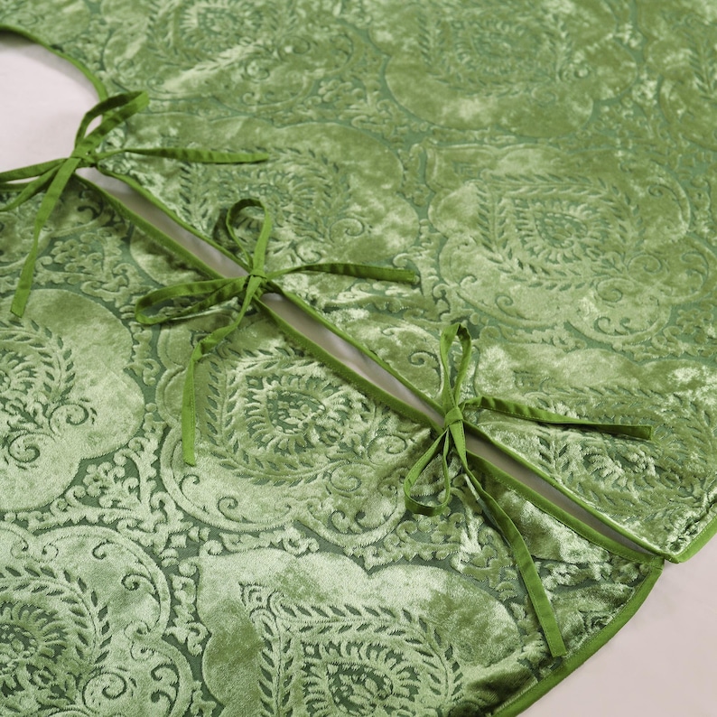 Puede incluir: Una falda de &aacute;rbol de Navidad de terciopelo verde con un patr&oacute;n floral detallado. La falda tiene un borde festoneado y se asegura con lazos de cinta verde. La tela tiene una textura suave y afelpada, y el dise&ntilde;o general es elegante y festivo.