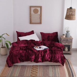 May include: A bed dressed with a deep red velvet duvet cover and matching cushions. A white pillow is in the centre. A patterned rug lies at the foot of the bed. A wooden bedside table and woven lamp are visible.