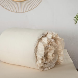 May include: A rolled-up, off-white comforter with a ruffled edge. The comforter is lying on a matching off-white bed sheet. The background includes a white wall, a decorative circular mirror, and a small wooden table.