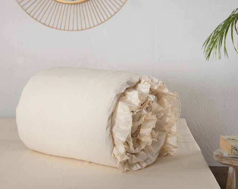 May include: A rolled-up, off-white comforter with a ruffled edge. The comforter is lying on a matching off-white bed sheet. The background includes a white wall, a decorative circular mirror, and a small wooden table.