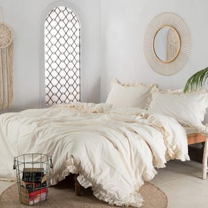 May include: A cream-colored bed with ruffled bedding and two matching pillows. A round mirror with a woven frame hangs above the bed. A macrame wall hanging and a wire basket filled with books are also visible.