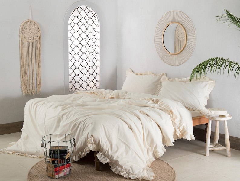 May include: A cream-colored bed with ruffled bedding and two matching pillows. A round mirror with a woven frame hangs above the bed. A macrame wall hanging and a wire basket filled with books are also visible.