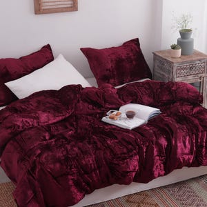 May include: A bed is made with a deep burgundy velvet comforter and matching pillowcases. A white pillow is also on the bed. A tray with a book and snacks sits on top of the comforter. A small wooden side table is next to the bed.