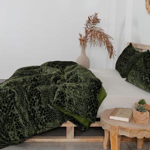 K&ouml;nnte beinhalten: Eine gr&uuml;ne Samtdecke und zwei passende Kissen mit einem reptilienartigen Muster werden auf einem Bett pr&auml;sentiert. Die Decke ist &uuml;ber einem Bettrahmen aus Holzpaletten drapiert. Ein kleiner Holztisch enth&auml;lt ein Buch und Topfpflanzen.