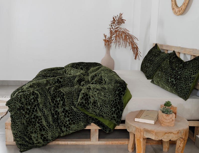 K&ouml;nnte beinhalten: Eine gr&uuml;ne Samtdecke und zwei passende Kissen mit einem reptilienartigen Muster werden auf einem Bett pr&auml;sentiert. Die Decke ist &uuml;ber einem Bettrahmen aus Holzpaletten drapiert. Ein kleiner Holztisch enth&auml;lt ein Buch und Topfpflanzen.