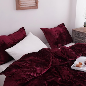 May include: A bed is dressed with a deep burgundy velvet duvet cover and matching cushions. A white pillow is also on the bed. A tray with tea and pastries sits on the duvet. The room is well-lit.