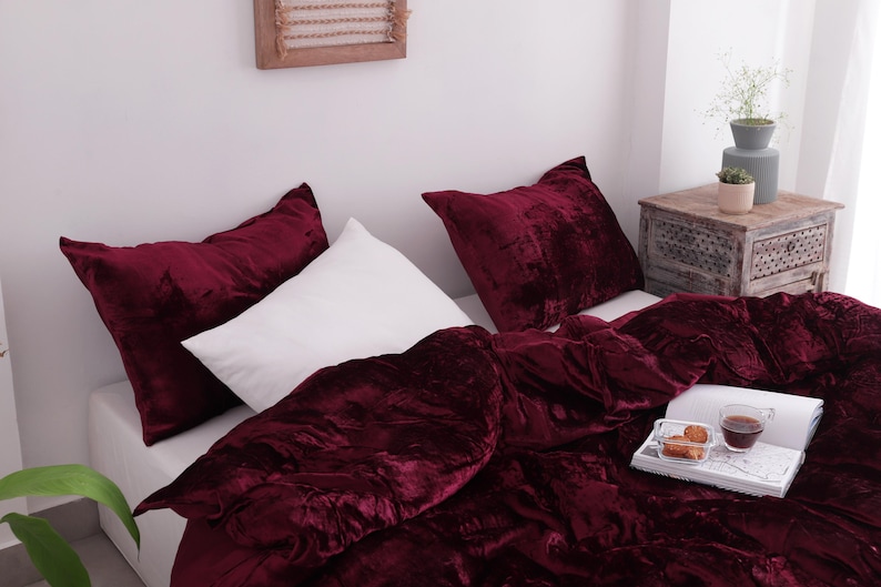 May include: A bed is dressed with a deep burgundy velvet duvet cover and matching cushions. A white pillow is also on the bed. A tray with tea and pastries sits on the duvet. The room is well-lit.
