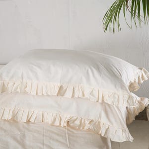May include: Two ivory-colored pillowcases with ruffled edges are stacked on top of each other. The pillowcases are made of a soft fabric and are part of a bedding set. The background is a plain white wall.