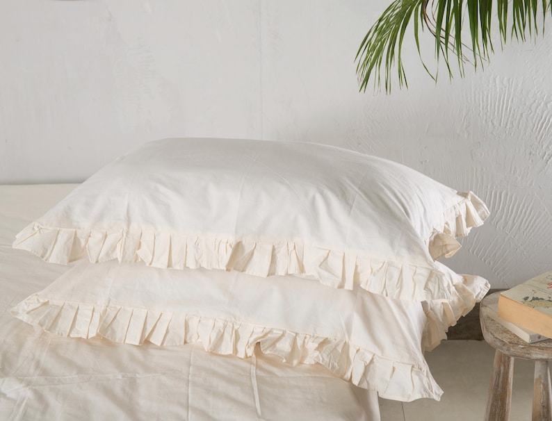 May include: Two ivory-colored pillowcases with ruffled edges are stacked on top of each other. The pillowcases are made of a soft fabric and are part of a bedding set. The background is a plain white wall.