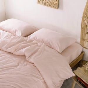 May include: A bed with light pink bedding, including a duvet and pillows. The bed is made up and ready for use. A wooden side table with a small vase and bowl is next to the bed. A woven pendant light hangs from the ceiling.