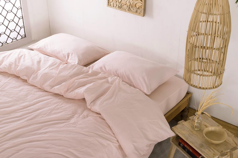 May include: A bed with light pink bedding, including a duvet and pillows. The bed is made up and ready for use. A wooden side table with a small vase and bowl is next to the bed. A woven pendant light hangs from the ceiling.
