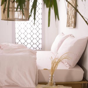 May include: A bedroom scene with a bed covered in light pink bedding and pillows. A wooden side table holds a glass vase with wheat stalks and a wooden bowl. A woven pendant light hangs from the ceiling.