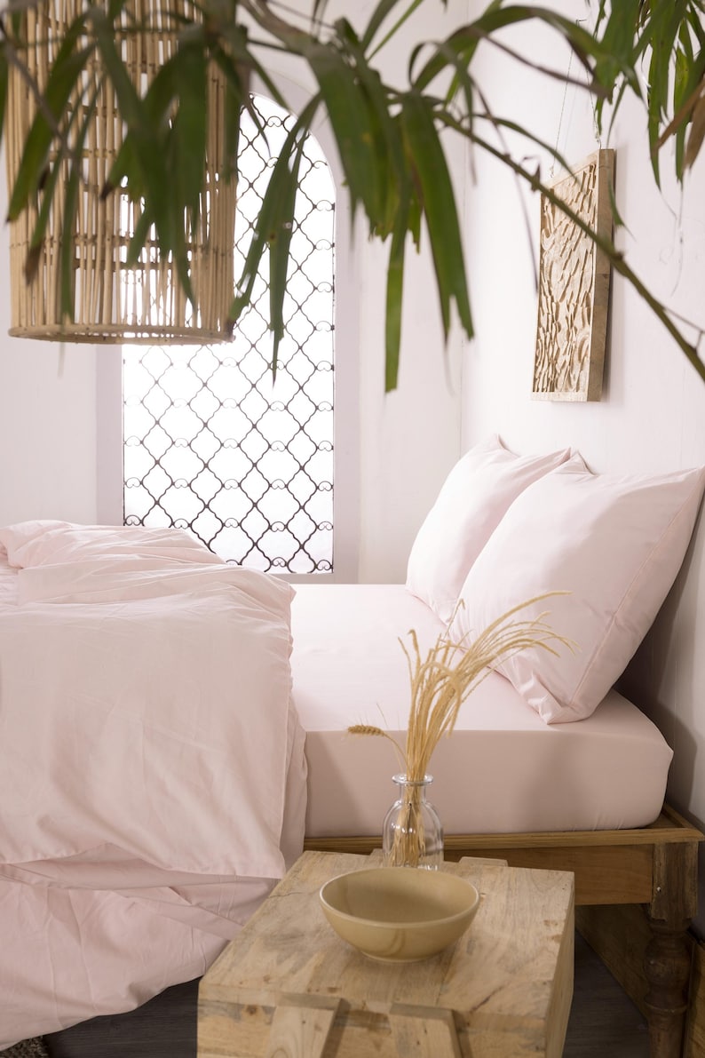 May include: A bedroom scene with a bed covered in light pink bedding and pillows. A wooden side table holds a glass vase with wheat stalks and a wooden bowl. A woven pendant light hangs from the ceiling.