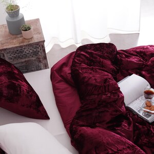 May include: A bed with a deep burgundy velvet comforter and matching pillow. A white pillow is also on the bed. A book, a small glass of liquid, and a plate of biscuits rest on the comforter. A small wooden side table is in the background.