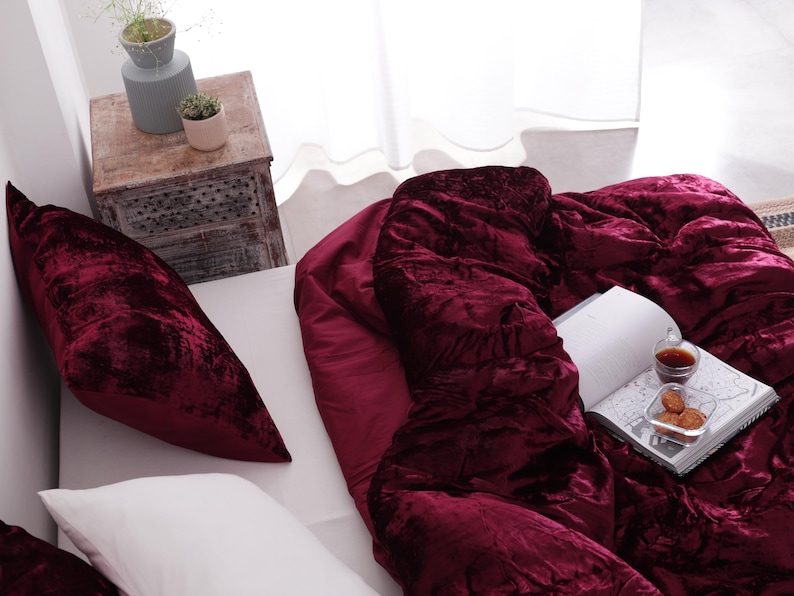 May include: A bed with a deep burgundy velvet comforter and matching pillow. A white pillow is also on the bed. A book, a small glass of liquid, and a plate of biscuits rest on the comforter. A small wooden side table is in the background.