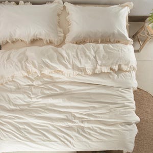 May include: An overhead shot of a bed with cream-colored bedding. Two ruffled pillowcases sit atop the bed, with a matching comforter and bedspread. The bedding is neatly arranged, creating a soft and inviting aesthetic. The overall theme is a cozy and comfortable bedroom.