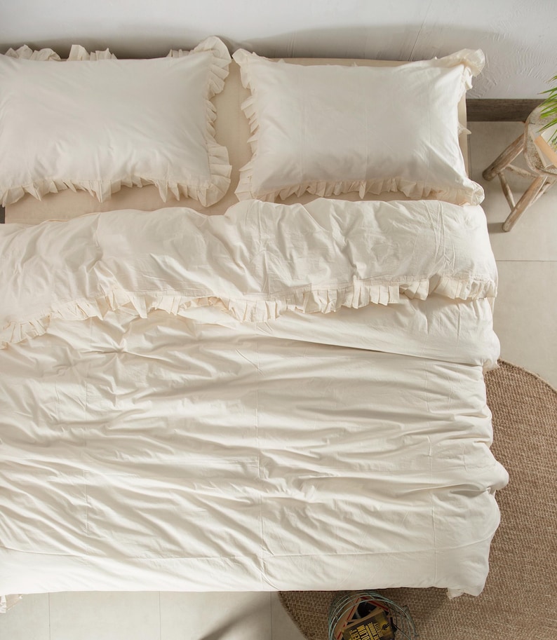 May include: An overhead shot of a bed with cream-colored bedding. Two ruffled pillowcases sit atop the bed, with a matching comforter and bedspread. The bedding is neatly arranged, creating a soft and inviting aesthetic. The overall theme is a cozy and comfortable bedroom.