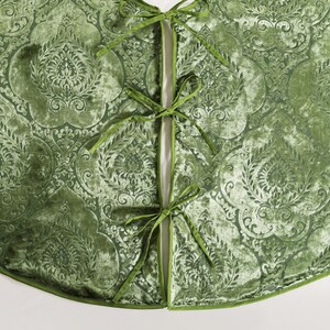 Puede incluir: Una falda de &aacute;rbol de Navidad de terciopelo verde con un patr&oacute;n en relieve ornamentado. La falda tiene una abertura dividida asegurada con lazos verdes. Los bordes est&aacute;n adornados con un borde verde a juego, creando una decoraci&oacute;n navide&ntilde;a festiva y elegante.