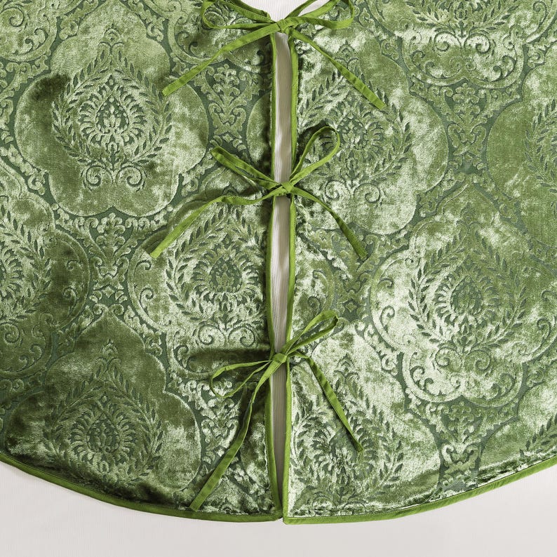 Puede incluir: Una falda de &aacute;rbol de Navidad de terciopelo verde con un patr&oacute;n en relieve ornamentado. La falda tiene una abertura dividida asegurada con lazos verdes. Los bordes est&aacute;n adornados con un borde verde a juego, creando una decoraci&oacute;n navide&ntilde;a festiva y elegante.