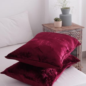 May include: Two burgundy velvet cushions stacked on a white bed. The cushions have a soft, textured appearance and a rectangular shape. The cushions are the focal point of the image, with a white pillow and a wooden bedside table in the background.