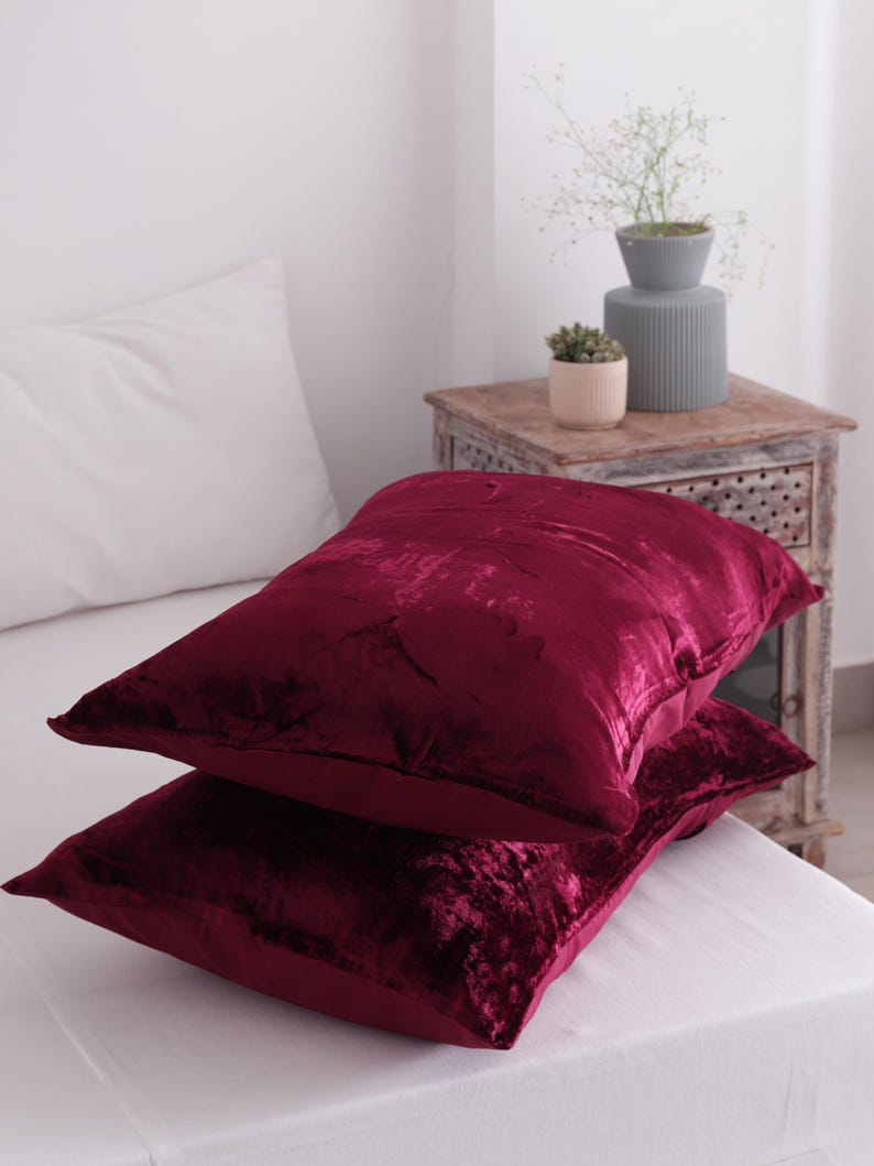 May include: Two burgundy velvet cushions stacked on a white bed. The cushions have a soft, textured appearance and a rectangular shape. The cushions are the focal point of the image, with a white pillow and a wooden bedside table in the background.