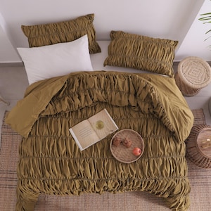 May include: Overhead view of a bed with olive green ruched bedding, including a duvet cover and two pillowcases. A white pillow is on the bed. A woven tray with fruit and a book are on the bed. A woven rug is under the bed.