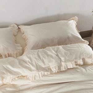 May include: Cream-colored bedding set featuring two ruffled pillowcases and a matching duvet cover. The bedding is made of a soft, light-colored fabric, creating a cozy and inviting atmosphere. The ruffled edges add a touch of elegance to the bedroom decor.