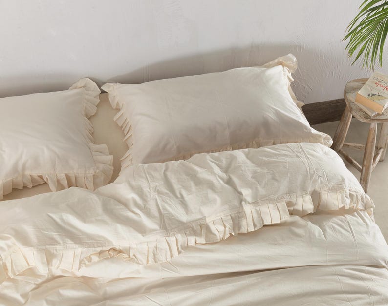 May include: Cream-colored bedding set featuring two ruffled pillowcases and a matching duvet cover. The bedding is made of a soft, light-colored fabric, creating a cozy and inviting atmosphere. The ruffled edges add a touch of elegance to the bedroom decor.