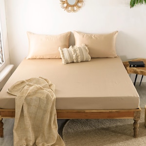 May include: A beige bed with matching pillowcases and a decorative pillow. A tan blanket is draped over the bed. The bed is made of wood and has a woven basket at the foot of the bed.