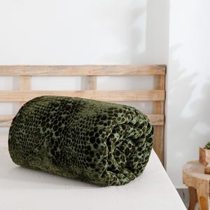 K&ouml;nnte beinhalten: Ein gerolltes, gr&uuml;nes Samt-Zierkissen mit einem dunklen, gefleckten Muster. Das Kissen liegt auf einem wei&szlig;en Bett, mit einem h&ouml;lzernen Kopfteil im Hintergrund. Die Textur des Kissens ist weich und pl&uuml;schig.