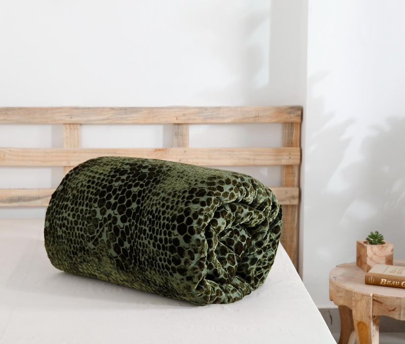 K&ouml;nnte beinhalten: Ein gerolltes, gr&uuml;nes Samt-Zierkissen mit einem dunklen, gefleckten Muster. Das Kissen liegt auf einem wei&szlig;en Bett, mit einem h&ouml;lzernen Kopfteil im Hintergrund. Die Textur des Kissens ist weich und pl&uuml;schig.