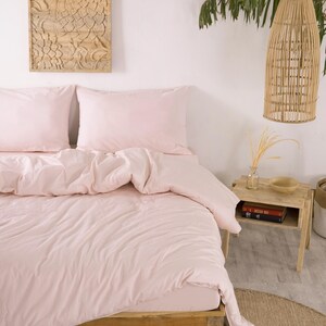 May include: A bedroom scene featuring a bed with light pink bedding. The bed is made of wood and has a matching duvet cover and pillowcases. A wooden carved wall hanging and a woven lamp shade are also visible.