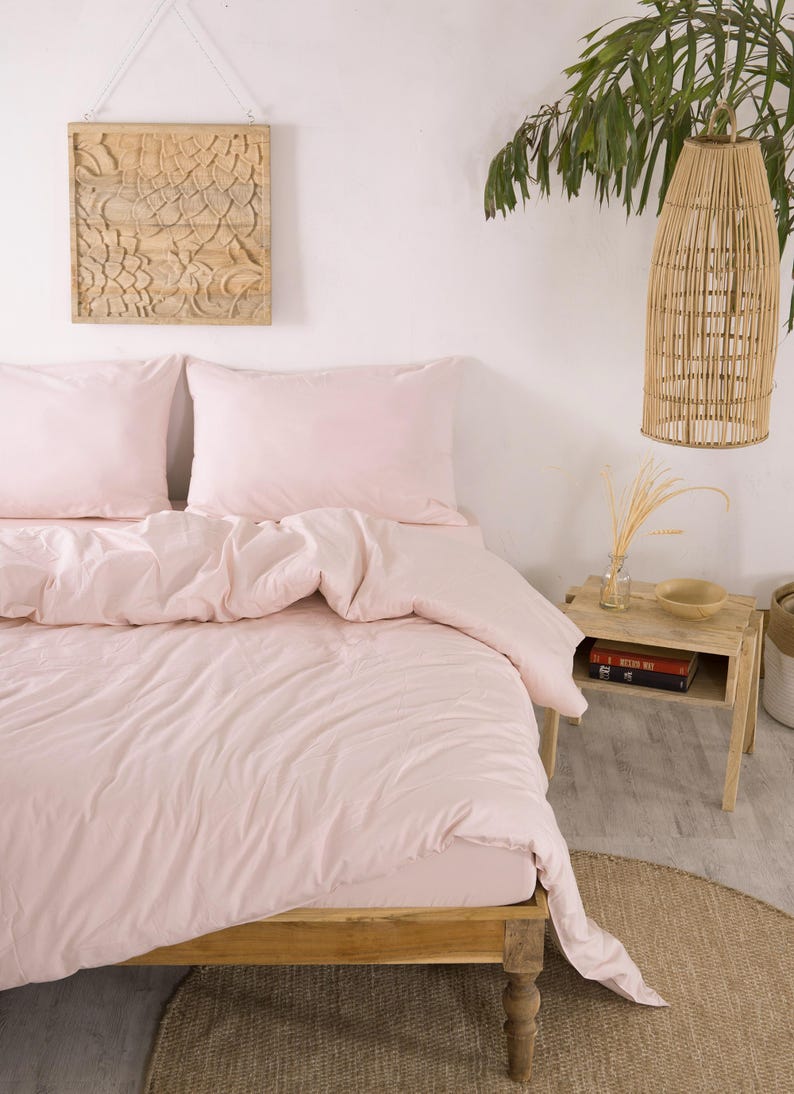 May include: A bedroom scene featuring a bed with light pink bedding. The bed is made of wood and has a matching duvet cover and pillowcases. A wooden carved wall hanging and a woven lamp shade are also visible.