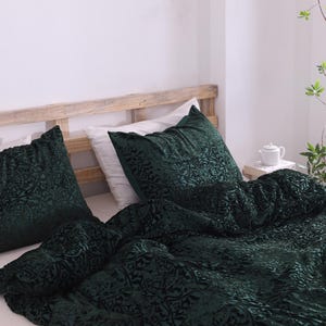 Könnte beinhalten: Ein Bett mit einer dunkelgrünen Samt-Bettdecke und passenden Kissenbezügen. Die Bettwäsche hat ein Blumenmuster. Das Bettgestell ist aus hellem Holz. Eine weiße Teekanne und eine Pflanze stehen auf einem Beistelltisch.