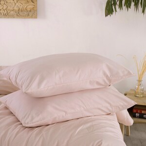 May include: Two light pink pillows stacked on a bed with matching bedding. The pillows are rectangular and appear soft. The bedding is a solid, light pink color, creating a cohesive and calming aesthetic.
