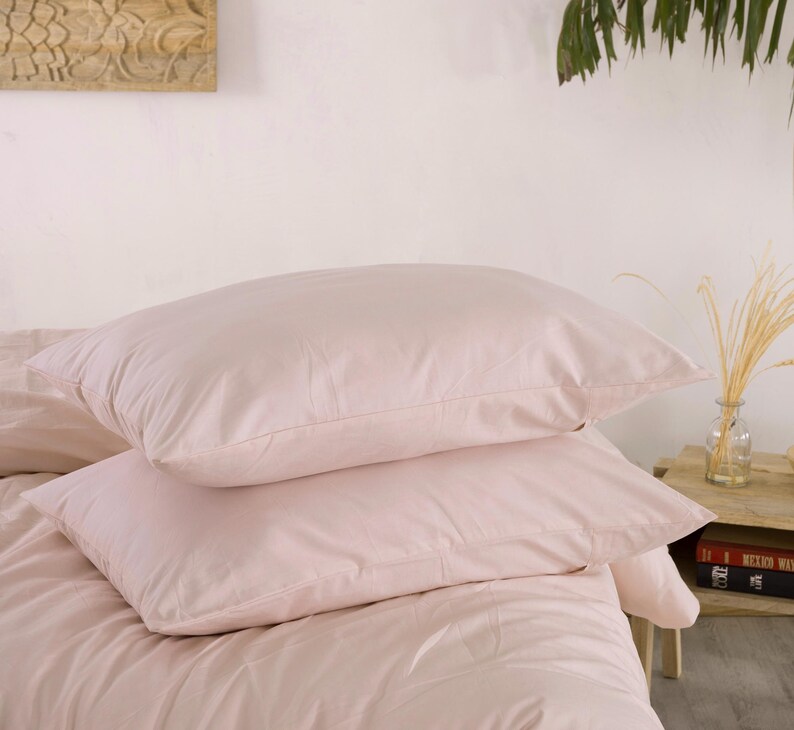 May include: Two light pink pillows stacked on a bed with matching bedding. The pillows are rectangular and appear soft. The bedding is a solid, light pink color, creating a cohesive and calming aesthetic.
