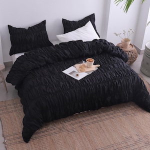 May include: A black textured bedspread and matching pillowcases are arranged on a bed. A white pillow is visible. A small wooden table holds a book and decorative items. A woven rug is on the floor.