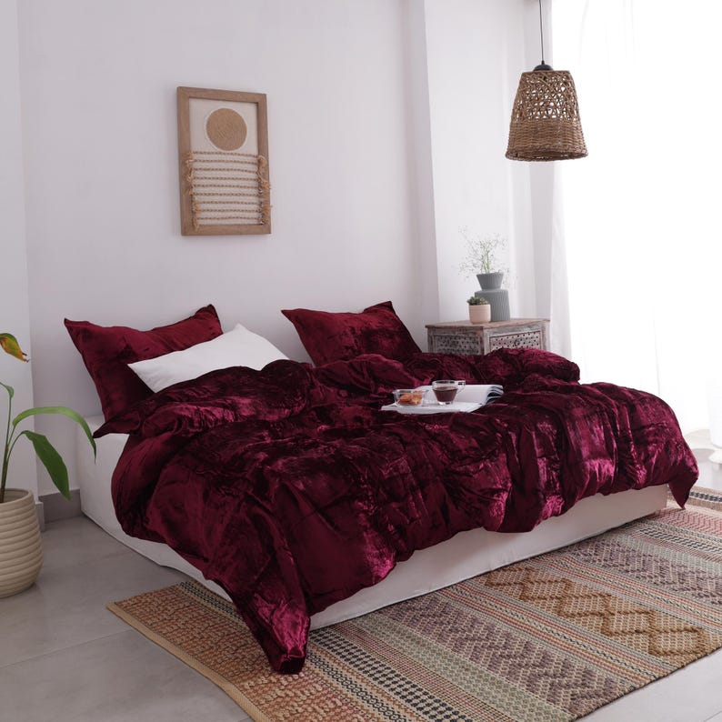 May include: A bed dressed with a deep burgundy velvet duvet cover and matching pillowcases. A white pillow is visible, along with a tray with cups and snacks. The bed is on a patterned rug.