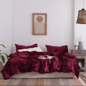 May include: A bedroom with a bed dressed in a deep red velvet duvet cover and pillows. A white pillow is placed on top. A woven pendant light hangs above the bed. A framed artwork and a bedside table are also visible.