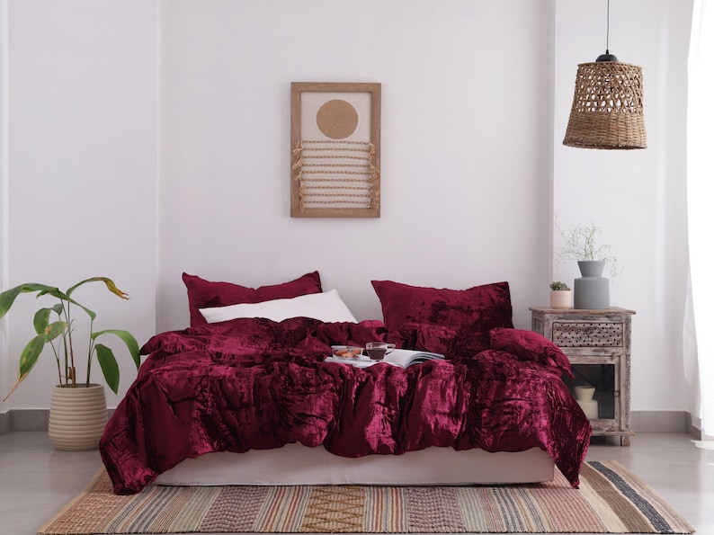 May include: A bedroom with a bed dressed in a deep red velvet duvet cover and pillows. A white pillow is placed on top. A woven pendant light hangs above the bed. A framed artwork and a bedside table are also visible.