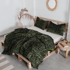 K&ouml;nnte beinhalten: Ein Bett mit einer gr&uuml;n-schwarz gemusterten Bettdecke und zwei passenden Kissen. Das Bettgestell ist aus Holz. Ein kleiner Nachttisch aus Holz enth&auml;lt ein Buch und kleine Topfpflanzen. Eine Vase mit getrockneten Pflanzen befindet sich links.