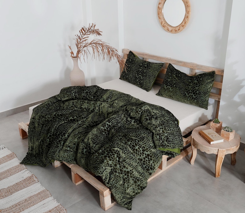 K&ouml;nnte beinhalten: Ein Bett mit einer gr&uuml;n-schwarz gemusterten Bettdecke und zwei passenden Kissen. Das Bettgestell ist aus Holz. Ein kleiner Nachttisch aus Holz enth&auml;lt ein Buch und kleine Topfpflanzen. Eine Vase mit getrockneten Pflanzen befindet sich links.