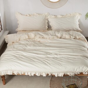 May include: Cream-colored bedding set with ruffled edges, including a duvet cover and two pillowcases. The bed is on a wooden frame, and the room has a neutral color palette with a round mirror and a small wooden stool.