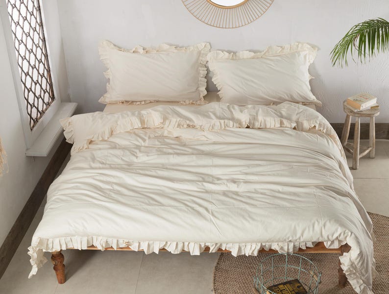May include: Cream-colored bedding set with ruffled edges, including a duvet cover and two pillowcases. The bed is on a wooden frame, and the room has a neutral color palette with a round mirror and a small wooden stool.