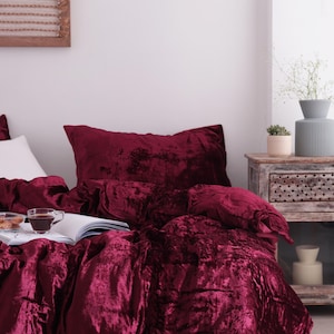 May include: A bed dressed with a deep red velvet duvet cover and pillow. A cup of coffee and a book rest on the bed. The room features a neutral colour scheme, a wooden bedside table, and decorative items.