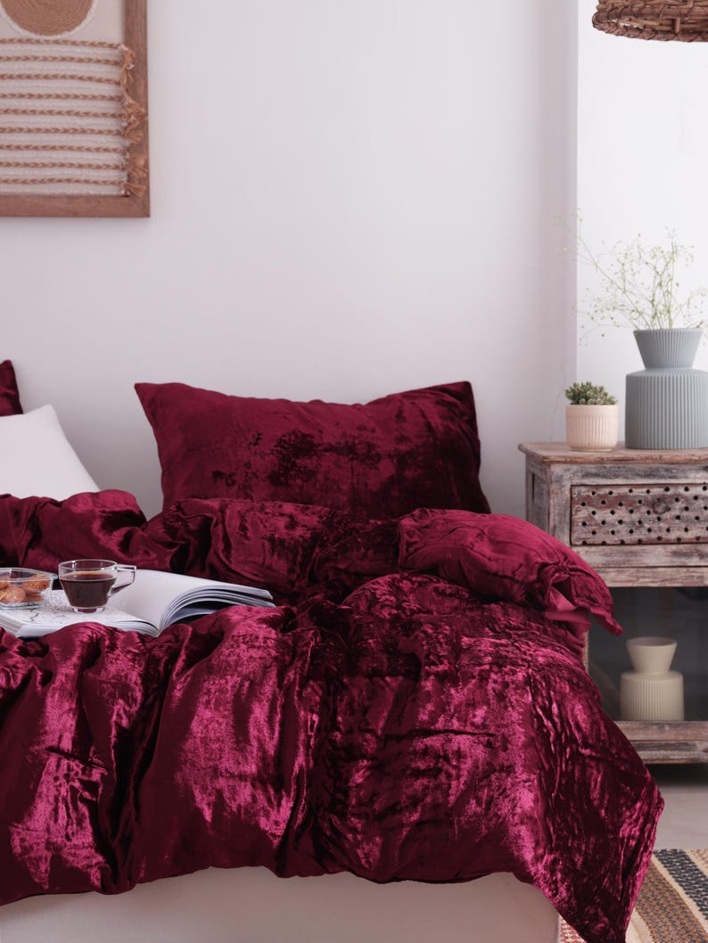 May include: A bed dressed with a deep red velvet duvet cover and pillow. A cup of coffee and a book rest on the bed. The room features a neutral colour scheme, a wooden bedside table, and decorative items.