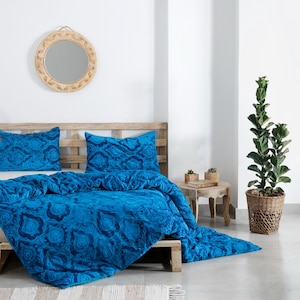 Könnte beinhalten: Ein Bett mit einem hölzernen Kopfteil und einem leuchtend blauen Bettdecken- und Kissensatz. Die Bettwäsche weist ein detailliertes, sich wiederholendes Muster auf. Über dem Bett hängt ein runder, gewebter Spiegel. Eine Topfpflanze und ein kleiner Beistelltisch sind ebenfalls zu sehen.
