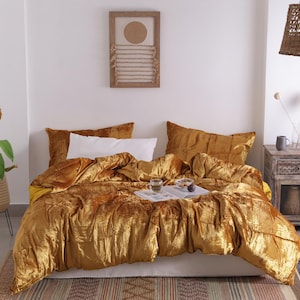 May include: A bed with a mustard-colored velvet comforter and matching pillows. A white pillow is visible. A woven pendant light hangs above a nightstand with a vase and a mug. A framed artwork and a potted plant are also in the room.